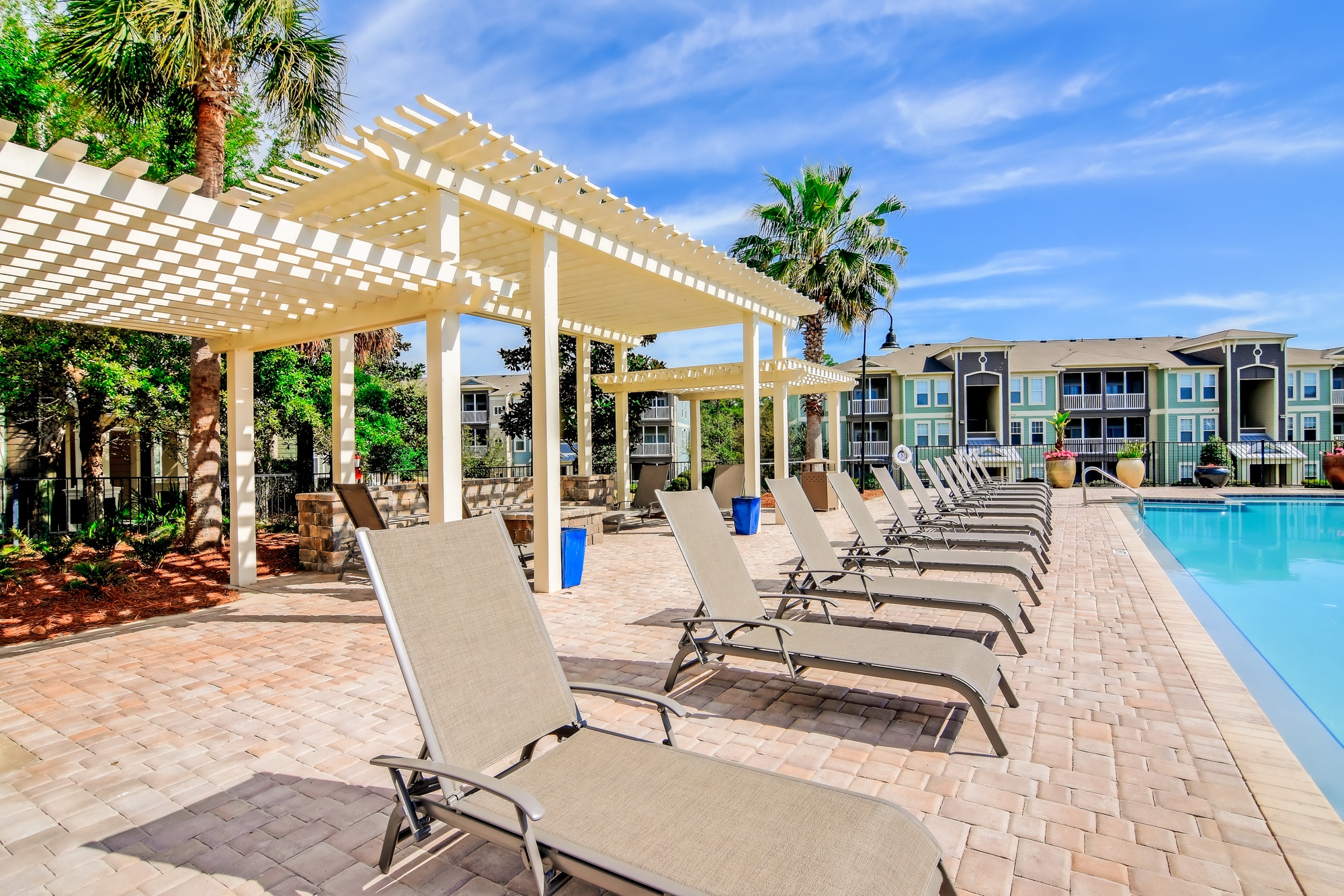 A row of sun loungers are set up under a white pergola by a pool.